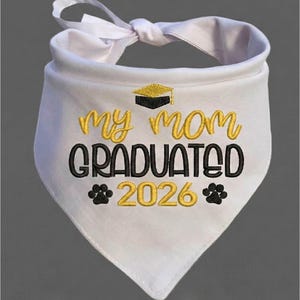 May include: White pet bandana with a satin ribbon tie. The bandana features embroidered text in gold and black that reads "My Mom Graduated 2026" with a graduation cap and paw prints.