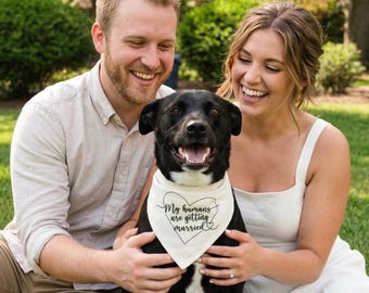 Peut inclure: Un chien noir portant un bandana blanc avec le texte "My humans are getting married" dans un motif en forme de cœur. Le chien est entouré de deux personnes souriantes, probablement lors d'une célébration de mariage ou de fiançailles.