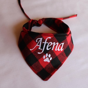 May include: Red and black plaid dog bandana with white embroidery. The bandana has the name "Afena" and a white paw print.