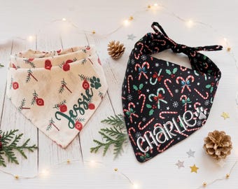 May include: Two triangular pet bandanas. One is cream-colored with a pattern of red ornaments, green pine needles, and the name Jessie in green script. The other is black with a pattern of candy canes, holly, and the name Charlie in white.
