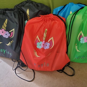 May include: Four drawstring bags with unicorn designs. The bags are black, red, blue, and green. The bags have the names "Kaylee", "Ayla", "Niki", and "Ayla" written on them.