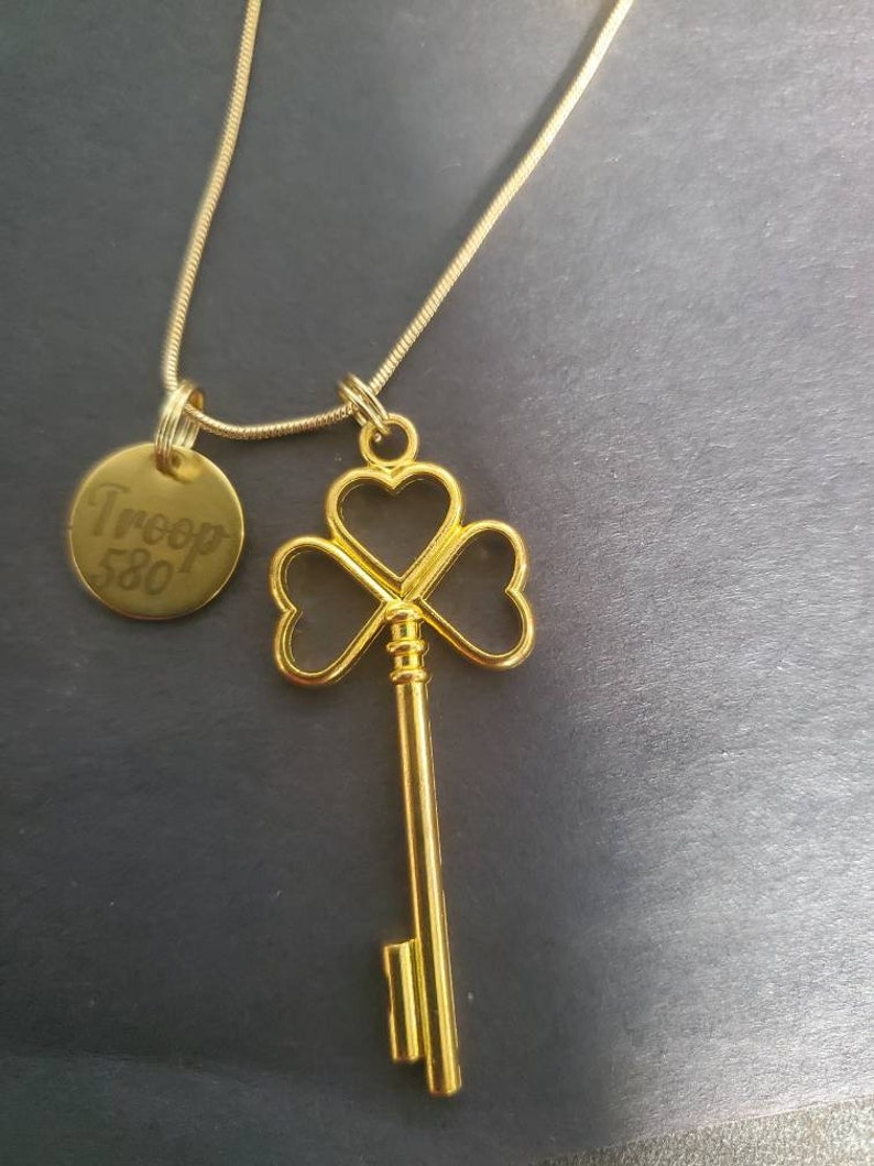 May include: A gold necklace with a key charm shaped like a four-leaf clover and a round tag that says "Troop 580".