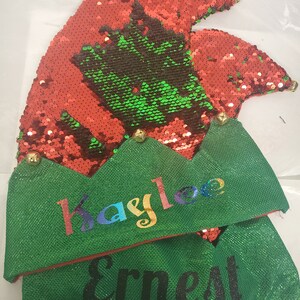 May include: Two green elf hats with gold bells and sequins. The top hat has a red sequin top and the name "Kaylee" written in rainbow colors. The bottom hat has the name "Ernest" written in black.