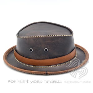 May include: A brown leather hat with a wide brim and a brown leather band. The hat has two metal eyelets on the front and is stitched with a light-colored thread. The hat is a classic style, suitable for outdoor wear.