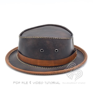 May include: A dark brown leather hat with a wide brim and a brown leather band. The hat has two small eyelets on the front and is stitched with a light-colored thread. The hat is a classic style, suitable for outdoor activities.