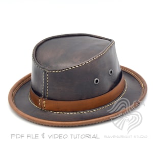 May include: A brown leather hat with a wide brim and a tan leather band. The hat has two small metal eyelets on the side and is stitched with cream-colored thread. The brim and band are a lighter shade of brown. The hat is a classic style.