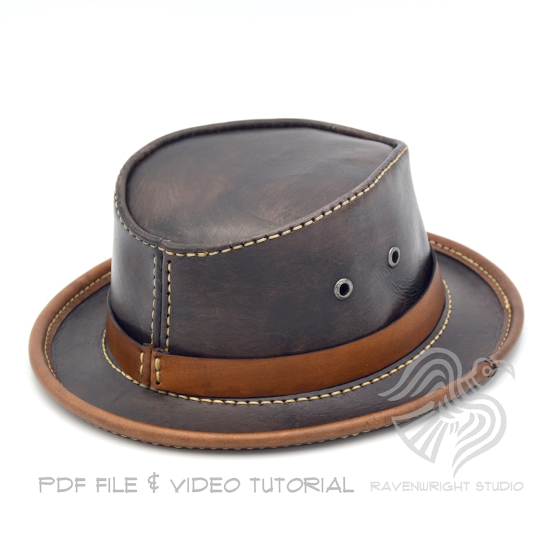 May include: A brown leather hat with a wide brim and a tan leather band. The hat has two small metal eyelets on the side and is stitched with cream-colored thread. The brim and band are a lighter shade of brown. The hat is a classic style.