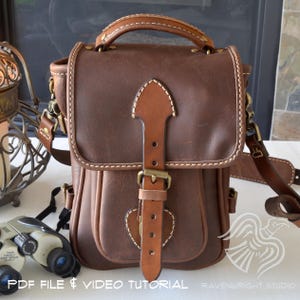 May include: A brown leather satchel with a top handle and adjustable shoulder strap. The bag features a flap closure with a leather strap and brass buckle. White stitching accents the edges. The text "PDF FILE & VIDEO TUTORIAL" is visible at the bottom.