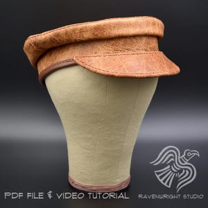 May include: A tan leather cap with a flat top and a short, curved brim. The cap is displayed on a mannequin head. The brim and the edge of the cap are trimmed with a darker brown leather. The text "PDF FILE & VIDEO TUTORIAL" is at the bottom.