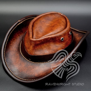 Può includere: Un cappello da cowboy in pelle marrone con una corona arrotondata e un'ampia tesa. Il cappello presenta una fascia marrone scuro attorno alla corona e cuciture colorate chiare. Il logo Ravenwright Studio è visibile.