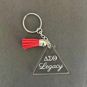 Könnte beinhalten: Transparenter Acryl-Schlüsselanhänger mit silbernem Schlüsselring und Kette. Der Schlüsselanhänger hat eine rote Quaste und eine dreieckige Form mit dem Text "ΔΣΘ Legacy" in weißer Schrift. Der Schlüsselanhänger befindet sich auf einem dunkelgrauen Hintergrund.
