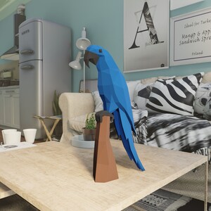 May include: A blue origami parrot perched on a brown wooden branch. The parrot is in a living room setting with a couch, coffee table, and a lamp.