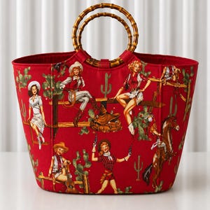 May include: A red tote bag featuring a repeating print of cowgirls, cacti, horses, and saddles. The bag has two circular bamboo handles attached to the top. The fabric displays various figures in western attire, including hats and boots.