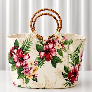 May include: A tote bag featuring a cream-colored base with a vibrant floral print of red hibiscus and yellow plumeria flowers surrounded by green palm leaves. The bag includes two circular bamboo handles attached to the top edge of the bag.