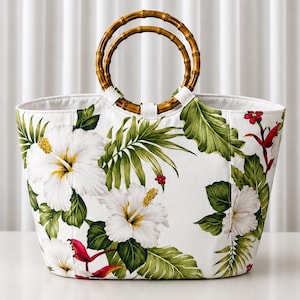 Peut inclure: Un sac fourre-tout blanc orné d'un imprimé tropical vif composé de grandes fleurs d'hibiscus blanches, de feuilles de palmier vertes et de petites touches rouges. Le sac est doté de deux anses circulaires en bambou fixées sur le dessus.
