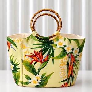 May include: A tote bag featuring a light yellow background with a vibrant tropical print of orange bird of paradise flowers, white plumeria blossoms, and green palm leaves. The bag includes two circular bamboo handles and a cream-colored interior.