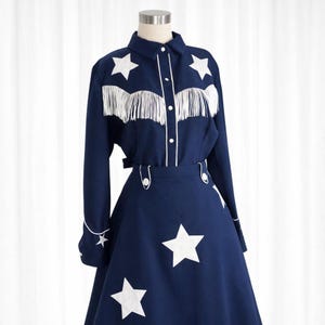 May include: A navy blue western-style outfit featuring a long-sleeved shirt and a skirt. The shirt has white fringe and star accents, with white piping. The skirt is adorned with large white stars. The outfit is displayed on a mannequin.