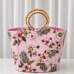May include: A tote bag featuring a light pink fabric base with a repeating floral pattern of hibiscus flowers in shades of pink, white, and yellow, accented by green palm leaves. The bag includes two circular bamboo handles at the top.