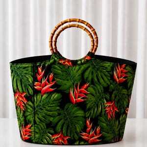 May include: A tote bag featuring a black background with a vibrant tropical print of green monstera leaves and bright orange-red heliconia flowers. The bag is equipped with two circular, segmented bamboo handles attached to the top opening.