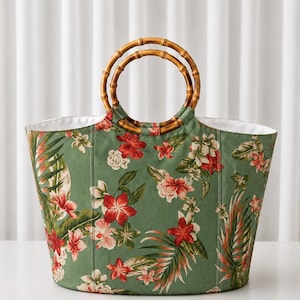 May include: A tote bag featuring a sage green fabric base with a vibrant floral print of red and white hibiscus flowers and green palm leaves. The bag includes two circular bamboo handles and a white interior lining, providing a structured shape.