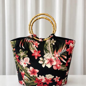 Puede incluir: Un bolso tote negro con un vibrante estampado floral tropical de flores rojas, rosas y blancas con hojas verdes. El bolso cuenta con dos asas circulares de bambú que están sujetas a la parte superior del cuerpo de tela.