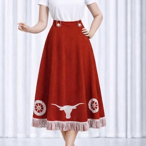 May include: A rust-coloured skirt featuring a white longhorn graphic and wheel designs near the hem. The skirt has a fringed bottom and white buttons at the waist. The skirt is paired with a white t-shirt.