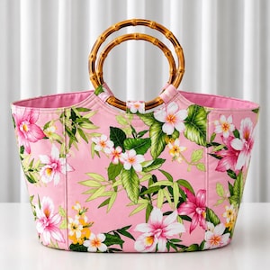 May include: A pink tote bag featuring a vibrant floral print with white, yellow, and magenta flowers and green leaves. The bag has two circular bamboo handles attached to the top, and the interior lining is a solid light pink fabric.