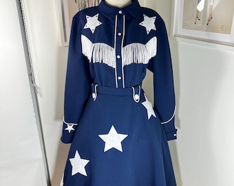 May include: A dark blue two-piece outfit consisting of a long-sleeved button-down shirt and a skirt. The shirt features white star appliqués, white piping, and white fringe across the chest. The skirt includes white star appliqués and white fringe trim.