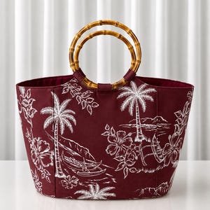 May include: A deep burgundy tote bag featuring a white line-art print of tropical palm trees, hibiscus flowers, and outrigger canoes. The bag is equipped with two circular bamboo handles attached to the top edge with matching fabric tabs.