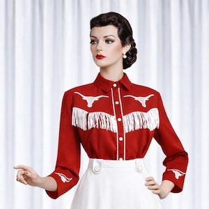 May include: A red long-sleeved shirt featuring white fringe and white embroidered longhorn designs. The shirt has white buttons and white trim. It is paired with a white skirt.