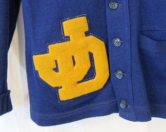 May include: A blue cardigan sweater with a yellow felt letter patch on the left side and a yellow felt letter patch on the right sleeve. The cardigan has a v-neck and five buttons down the front. The label on the cardigan reads "Deauville".