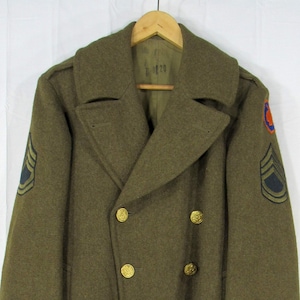 May include: Olive green military overcoat with a double-breasted design and gold-colored buttons. The coat features shoulder insignia and a notched collar. The coat is hanging on a wooden hanger.