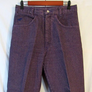May include: A pair of purple denim jeans hanging on a wooden hanger. The jeans have a classic five-pocket design with a button and zipper closure. A small blue embroidered design is visible on the front pocket. The jeans are made of a sturdy denim material.