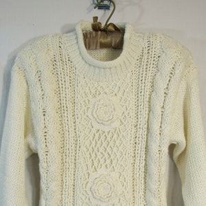 May include: Cream-colored, hand-knitted sweater with a round neckline and long sleeves. The sweater features three prominent floral designs and intricate cable and lattice patterns. The knitwear is displayed on a hanger.