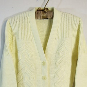 May include: A pale yellow cardigan sweater with a V-neck, button closure, and two front pockets. The sweater features a cable knit design on the front panels and ribbed detailing on the shoulders and cuffs. The sweater is hanging on a wooden hanger.