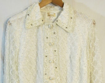 May include: Ivory-colored, long-sleeved blouse with a collared neckline. The blouse is adorned with small, shimmering beads and a diamond-patterned design. The cuffs and collar are also embellished with beads. The blouse is hanging on a wooden hanger.
