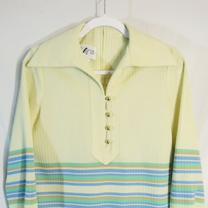 May include: A pale yellow long-sleeved shirt with a striped pattern in blue, green, and brown. The shirt has a collared neckline and a button closure. The shirt has a drawstring at the bottom.