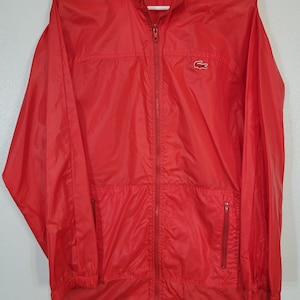 May include: A bright red, full-zip jacket with the "ALLIGATOR LACOSTE" logo on the collar. The jacket has two front pockets with zippers and the Lacoste crocodile logo on the chest. The jacket is made of a lightweight material.