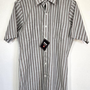 May include: Short-sleeved button-down shirt with vertical stripes in brown, white, and red. The shirt has a classic collar, two chest pockets, and a tag that reads "Arrow". The shirt is hanging on a hanger.