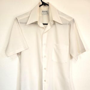 May include: Off-white short-sleeved shirt with a pointed collar and a chest pocket. The shirt has a button-down front and the brand name "Century" is visible on the label.