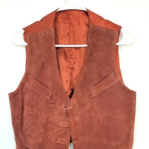 May include: A rust-colored suede vest with a V-neck and four buttons down the front. The vest has two front pockets and is hanging on a white hanger. The interior lining is a darker shade of rust.
