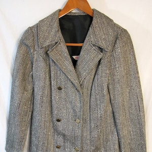 May include: A double-breasted blazer in a black and white herringbone pattern. The jacket has a notched collar, three gold-colored buttons, and a black lining. The blazer is hanging on a wooden hanger.