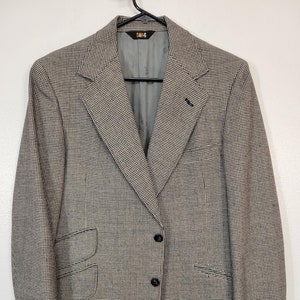 May include: A houndstooth pattern blazer in shades of tan and black, featuring a two-button closure and a notched lapel. The blazer has a classic design with a tailored fit, and three buttons on each cuff. The interior lining is gray.
