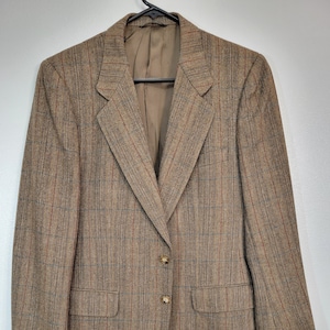 May include: A brown blazer with a plaid pattern. The blazer has two buttons, two front pockets, and a notched lapel. The plaid pattern includes red and blue lines. The blazer is hanging on a black hanger.