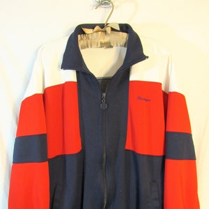 May include: A vintage track jacket in a color block design. The jacket features a white upper chest, red sleeves, and a navy blue center panel. It has a full zipper, a stand-up collar, and a small logo on the chest. The cuffs and waistband are ribbed.