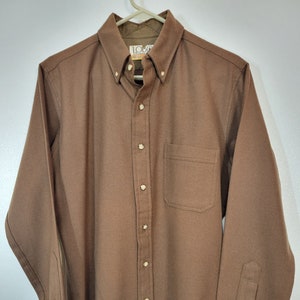May include: A brown button-down shirt with a button-down collar and a single chest pocket. The shirt has long sleeves with buttoned cuffs and is hanging on a white hanger. The shirt has the brand name "LOBO" on the collar.