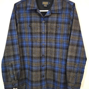 K&ouml;nnte beinhalten: Ein blaues, schwarzes und graues kariertes Button-Down-Hemd. Das Hemd hat lange &Auml;rmel, zwei Brusttaschen und einen Kragen. Das Pendleton-Markenetikett ist auf der Innenseite des Kragens sichtbar. Das Hemd h&auml;ngt an einem wei&szlig;en Kleiderb&uuml;gel.