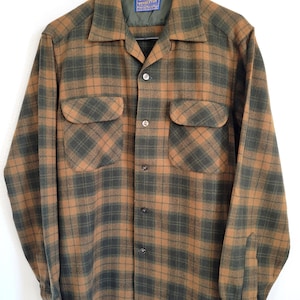 Pode incluir: Uma camisa de manga comprida com bot&otilde;es, com um padr&atilde;o xadrez castanho e verde. A camisa tem dois bolsos no peito e uma gola. Est&aacute; pendurada num cabide preto sobre um fundo branco.