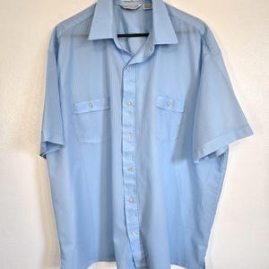 May include: Light blue short-sleeve shirt with vertical stripes, two chest pockets, and a button-down front. The shirt is hanging on a black hanger against a white wall. The shirt has a classic collar.