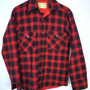 May include: Red and black plaid flannel shirt with two chest pockets and a collared neckline. The long-sleeved shirt is button-down and hangs from a black hanger. The shirt has a red lining.